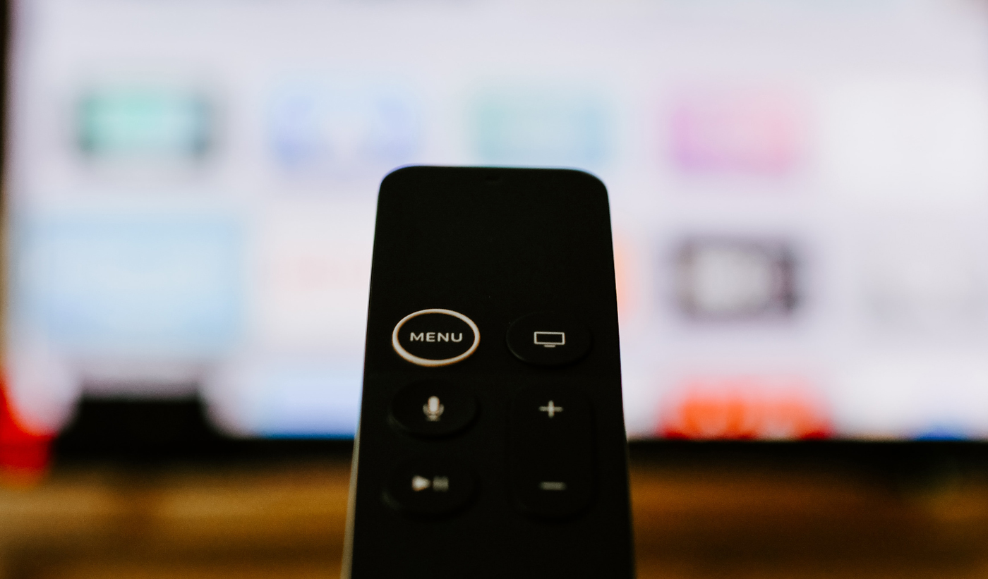 An Apple TV remote held up in-front of a TV with the Apple TV home page on