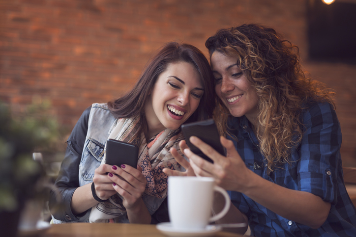 Two girls sitting in a cafe looking and laughing at their mobile phones