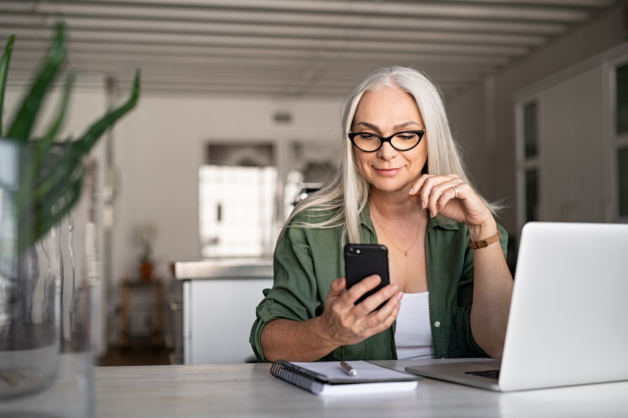 A woman sitting at a desk with a laptop and notepad on it using her mobile phone