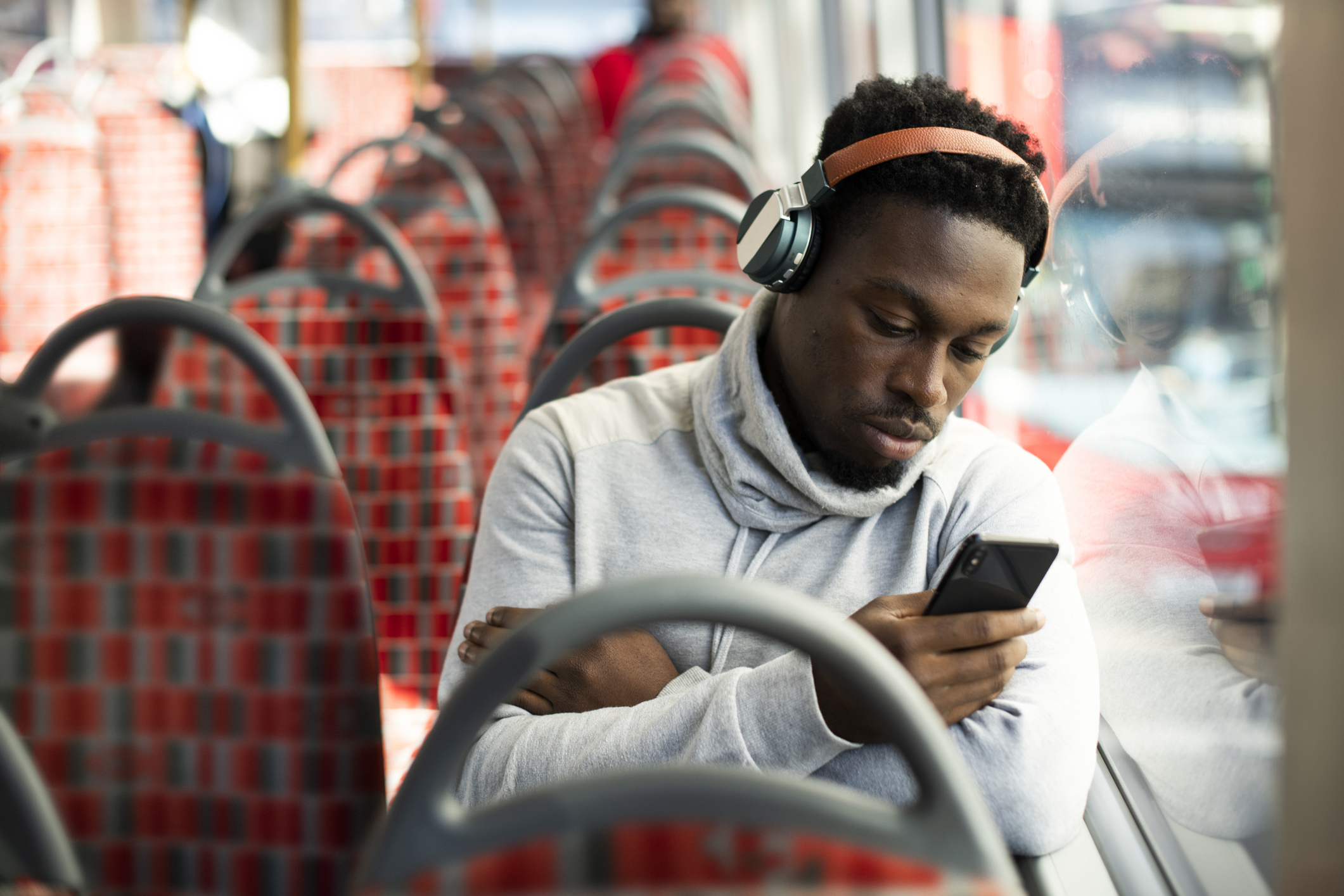 A man sitting on a bus listening to music on headphones whilst using his mobile phone