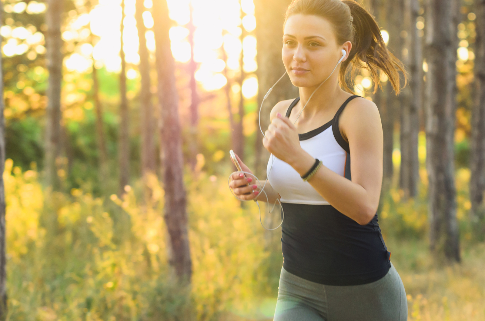 A woman running in a woodland area at sunset with headphones i  whilst holding her phone