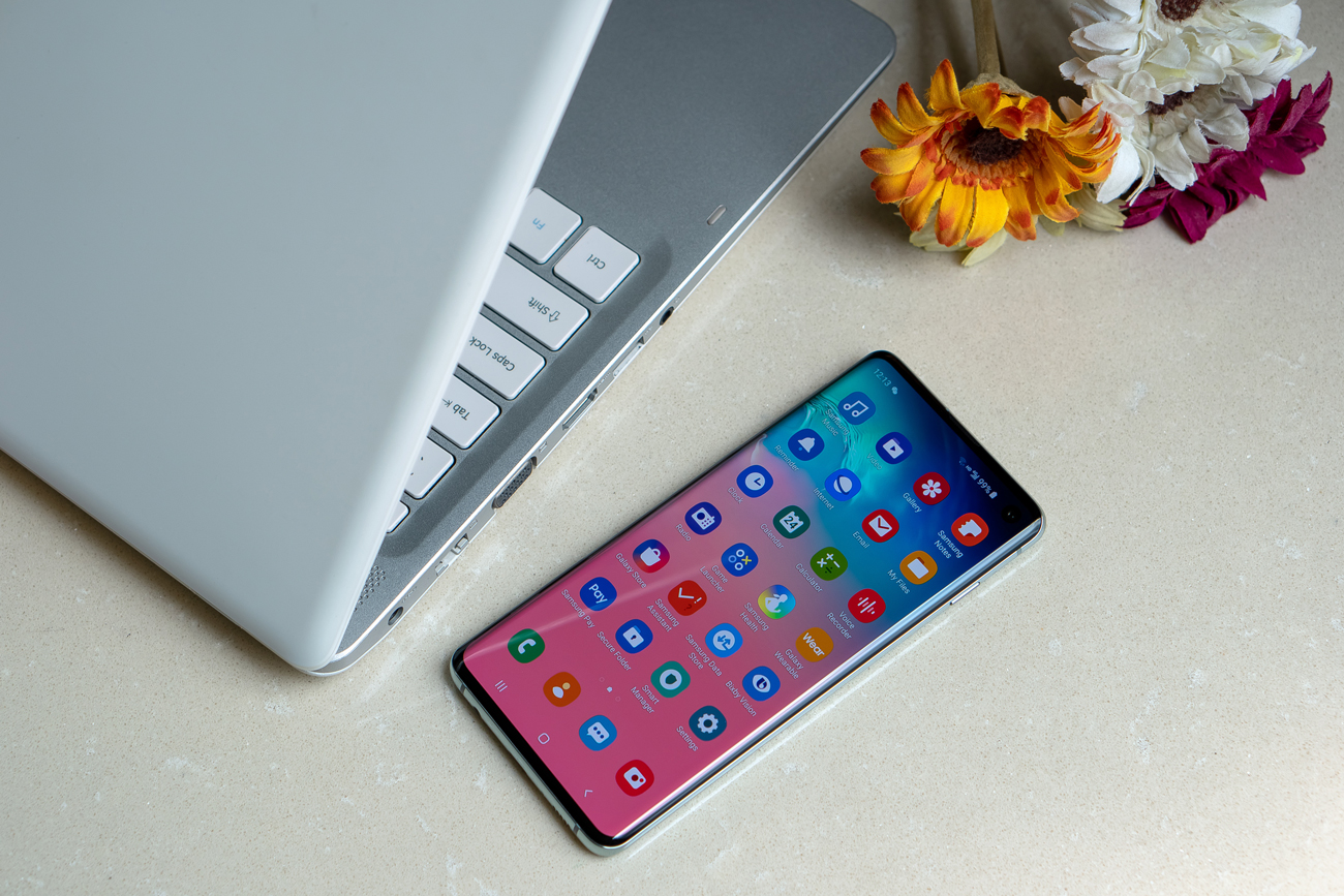 A Samsung Galaxy S10 on a table next to a laptop and flowers